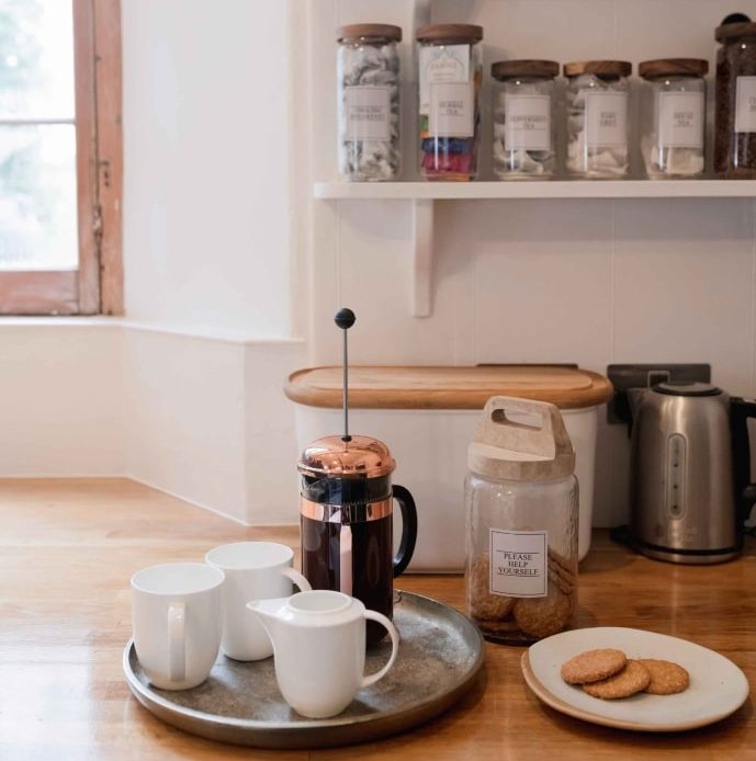 A tray with mugs and a cafetiere of local supplier coffee sits on a wooden counter next to a plate of biscuits. The shelves above are lined with teas.