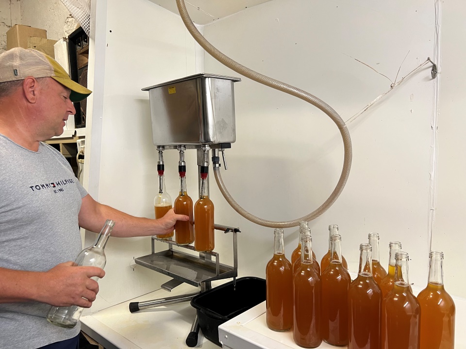A man attaches glass bottles to a metal machine with nozzles filling the bottles with apple juice.