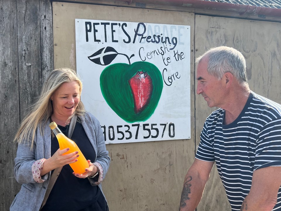 A woman holds a glass bottle of apple juice and smiles, and a man in a striped t shirt looks at it. There is a hand painted sign behind, reading Pete's Pressing with a phone number