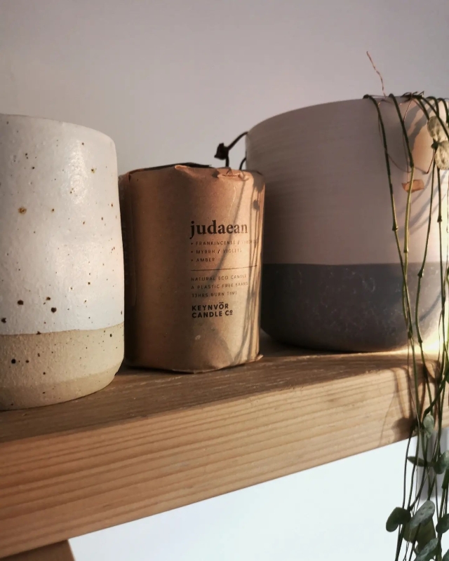 A candle wrapped in paper packaging between two plant pots on a shelf.