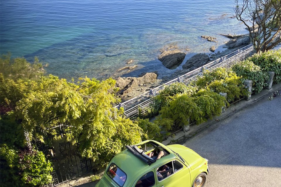 A small green car with a sun roof drives next to the sea front on a honeymoon in Cornwall