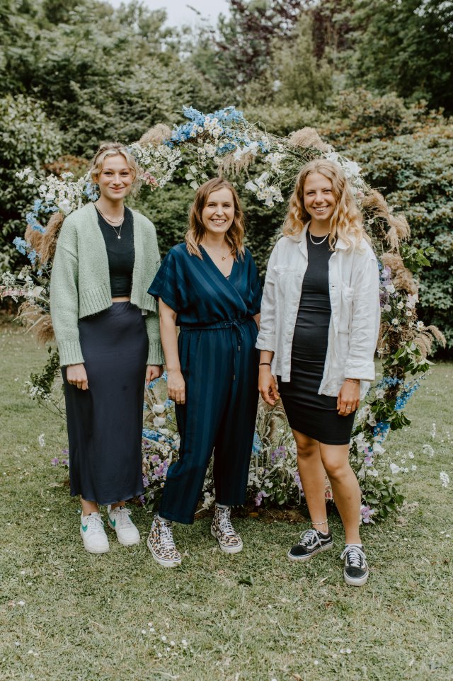 Floral arch set up in a garden with three women stood side by side and smiling.