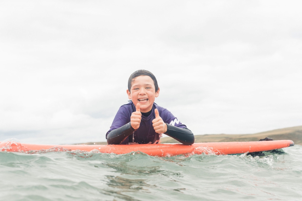 A child does thumbs up, leaning on a red surf board in the sea