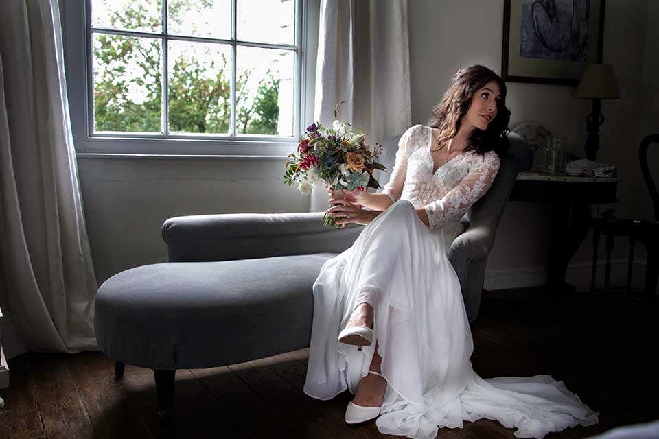 elegant bride in wedding dress sat on grey chaise lounge in bridal suite